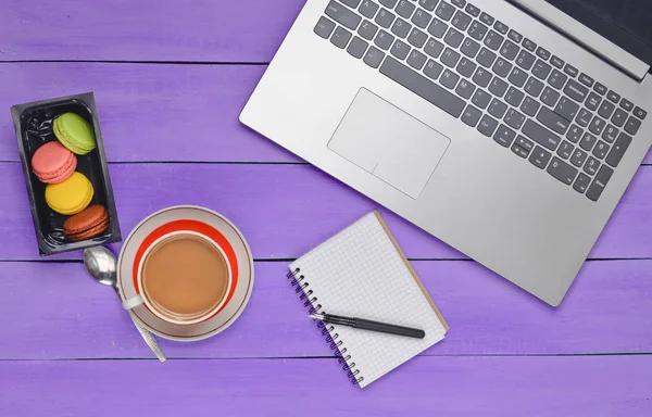 Laptop, colored macaroons cookies, cup of coffee, notebook with a pen on a blue wood table. Top view. Flat la