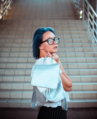 Gümüş kumaş ve kamera önünde poz ile bir sırt çantası elinde tutan bir çekici mavi saçlı öğrenci kız portresi. Sokak moda.