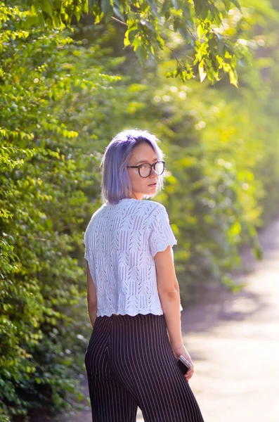 Street fashion. Stylish look. Blue-haired hipster girl in glasses posing in front of camera in park.