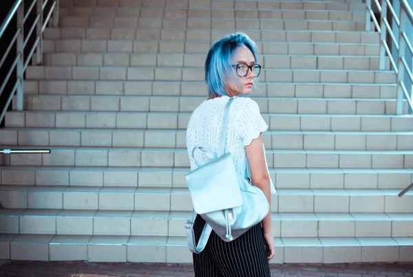 Atypical beauty attractive blue-haired girl with a backpack of silver fabric plans on the background of steps.
