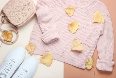 Bej arka planda kadın giyim ve aksesuarları. Moda kazak, spor ayakkabıları, deri çanta düşmüş Sarımlı yaprakları arasında. Sonbahar dolabı. üst görünüm