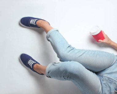 Kot pantolon ve spor ayakkabıları kız beyaz bir arka planda oturur ve kahve fincan tutar. Üst görünüm