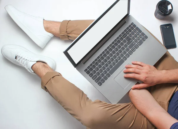 Fragment of male legs in beige pants and white sneakers sitting on white. Man using modern laptop. Online worker, freelancing, work at home, man working. top view on white backgroun