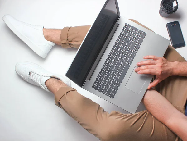 Fragment of male legs in beige pants and white sneakers sitting on white. Man using modern laptop. Online worker, freelancing, work at home, man working. top view on white backgroun