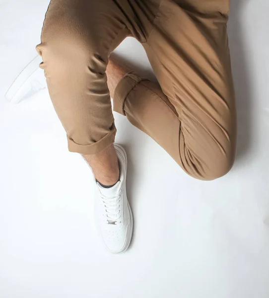 Fragment of male legs in beige trousers and white sneakers sits on a white background. Top view. Relaxatio