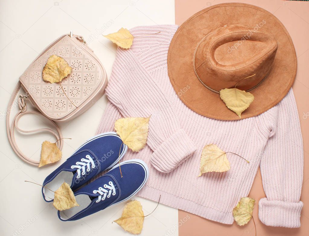 Women's clothing and accessories on a beige background. Fashionable sweater, sneakers, felt hat, leather bag among the fallen yellowed leaves. Autumn wardrobe. top view