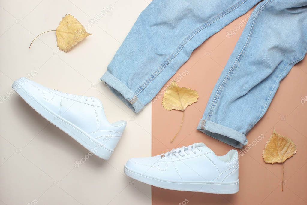 Jeans and white sneakers among the fallen leaves on a beige brown paper background. Minimalism, top view