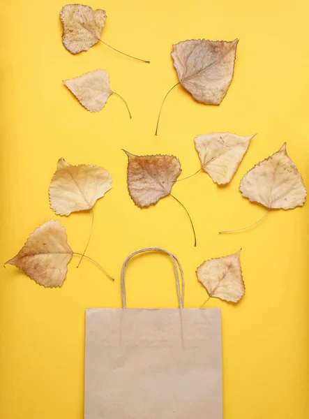 Paper bag for shopping, fallen autumn leaves on yellow background. Autumn shopping, sale, Top view, minimalism