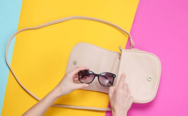 Kadın el kağıt renkli arka plan üzerinde deri çanta moda güneş gözlüğü alır. Üst görünüm, düz yatıyordu, minimalizm
