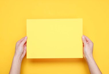 Kadın elleri sarı arka plan üzerinde sarı kağıt levha tutan. Üst te. Mockup, kopyalama alanı, minimalizm