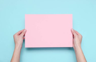 Kadın elleri mavi arka plan üzerinde pembe kağıt levha tutan. Üst te. Mockup, kopyalama alanı, minimalizm