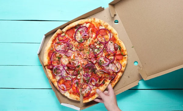 The hand takes a piece of pizza in box on blue concrete table. Top vie