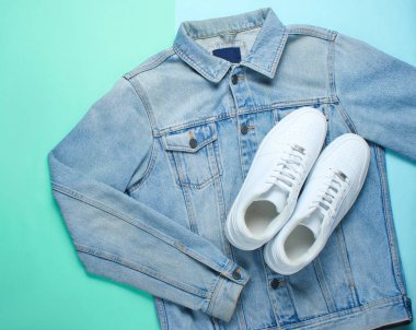 Düz bir pastel mavi arka planda bir denim ceket ile stil beyaz hipster Sneakers yatıyordu. Retro kültür, üst görünüm