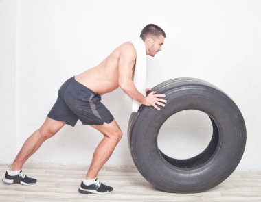 Young man athlete with a white towel rolls a big wheel for training crossfit against the background of the wall