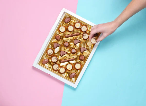 Girl takes a chocolate candy from a box of chocolates with a golden tray on blue pink pastel background. Top view, minimalis