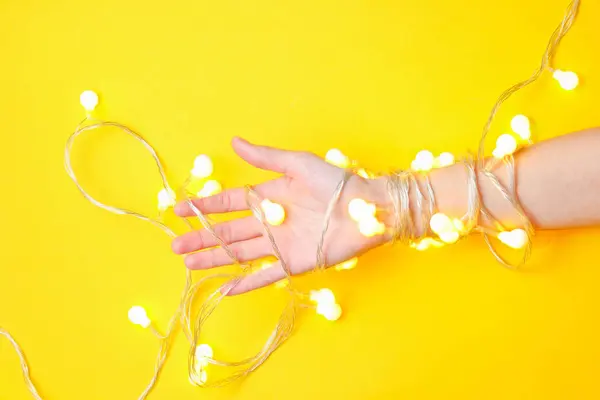 Minimalism trend. Female hand wrapped with bright luminous garlands of light bulbs on a yellow background. Top view