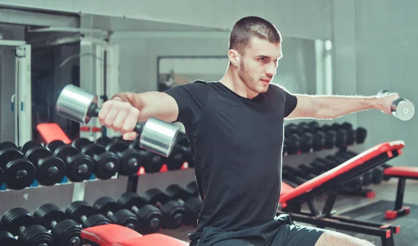 Atletik genç adam omuzlarını egzersiz ve spor salonunda dumbbells çifti ile yan yükseltir yapıyor