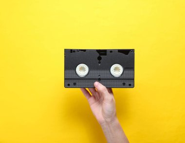 Kadın el sarı arka plan üzerinde video kaset tutar. Retro tarzı, pop kültürü, minimalizm, üst görünümü