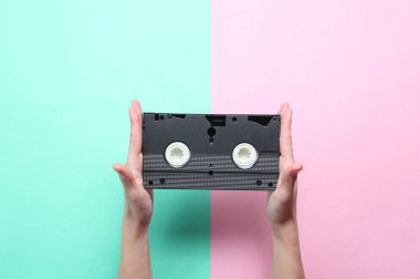 Kadın elleri pembe mavi pastel arka plan üzerinde video kaset tutar. Retro tarzı, pop kültürü, minimalizm, üst görünümü