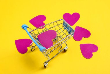 Mini alışveriş arabası, sarı arka plan üzerinde dekoratif kalpler. Kavramsal fotoğraf