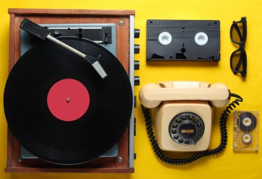 Sarı arka plandaki eski moda nesneler. Retro tarzı, 80'li, popüler kültür. Üst te. Vinil çalar, döner telefon, video, ses bandı