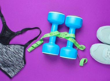 Fitness konsepti. Kilo kaybı. Spor sutyeni, ayakkabılar, dambıllar, mor arka planda hükümdar. Üst manzara, düz uzanma