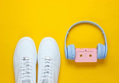 Retro tarzı. 80 'ler. Popüler kültür. Minimalizm. Ses kaseti olan kulaklıklar, sarı arka planda beyaz spor ayakkabılar. Üst görünüm
