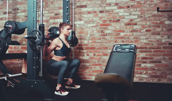 Gergin sportif kız çanları tuğla duvara karşı Smith makinede halter ile çömelme egzersiz