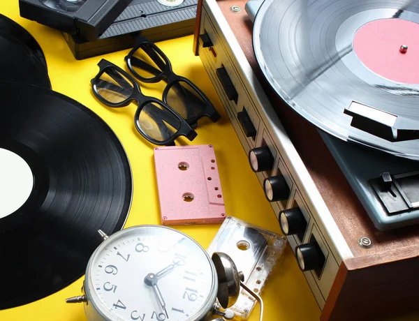 Vintage attributes, 80s media. Vinyl player, video cassettes, audio cassettes, records, radio, vintage alarm clock on a yellow background.