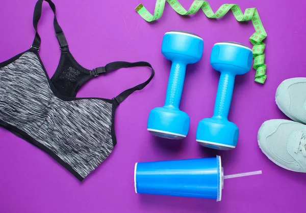 Fitness concept. Weight loss. Sports bra,shoes, dumbbells, ruller,cup on purple background. Top view, flat lay