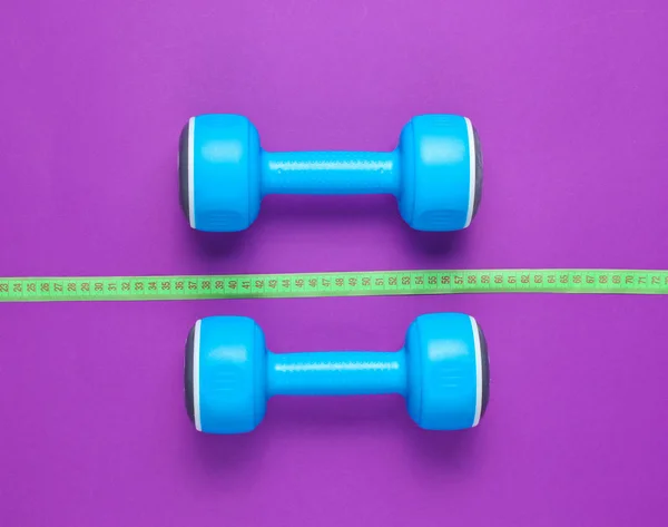 The concept of losing weight. Blue plastic dumbbells, ruler on purple background. Sport, fitness. Top view. Flat lay