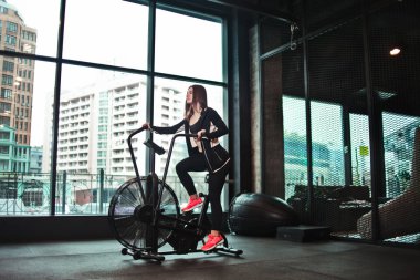 Kardiyo eğitimi. Spor salonunda egzersiz bisikleti kullanan genç sportif kadın. Fitness kız karanlık spor salonu ön pencerede kardiyo egzersiz için hava bisikleti kullanarak