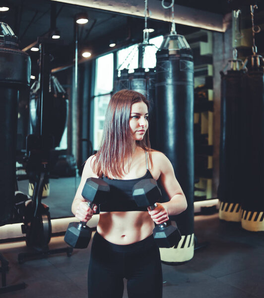Attractive athletic girl in sportswear holds dumbbells in her hands and trains biceps in boxing gym.