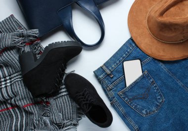 Üst düzey moda kıyafetler, ayakkabılar, gri arka planda aksesuarlar. Kot etek, şapka, fular, siyah botlar, deri çanta, arka cebinde akıllı telefon.