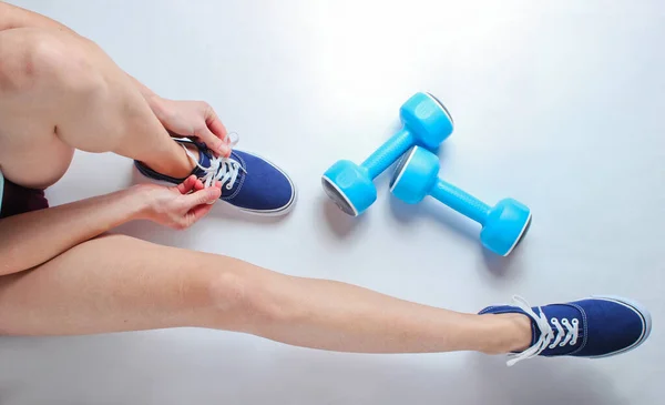 Legs of young sports woman, plastic dumbbells. Woman tying shoelace of sports shoes on a white background. Top view. Minimalism