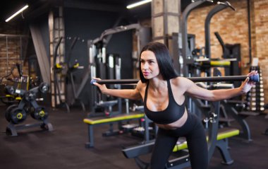 Çekici esmer sporcu spor salonunda vücudunu eğerek omuzlarındaki çıtayla jimnastik yapıyor.