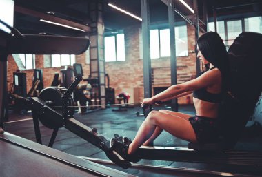 Simülasyon kürek egzersiz makinesinin üzerinde spor kıyafetleri içinde kasıtlı olarak oturan esmer. Fonksiyonel eğitim