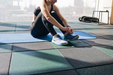 Spor ayakkabılarının bağcıklarını bağlayan kadın spor salonundaki paspasın üzerinde oturuyor.