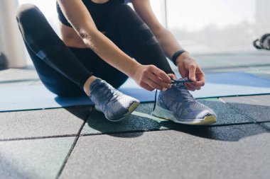 Spor ayakkabılarının bağcıklarını bağlayan kadın spor salonundaki paspasın üzerinde oturuyor.