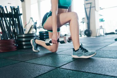 Spor salonunda elinde dambıllarla bacaklarına hücum eden genç fitness kızı..