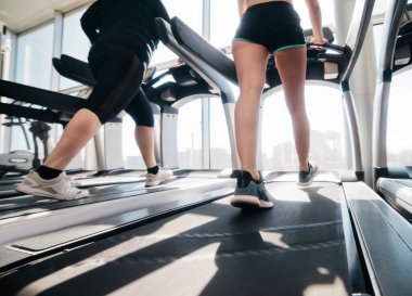 Kadınlar spor giyer ve spor ayakkabıları giyer. Spor salonunda pencereye karşı koşu bandında koşarlar.