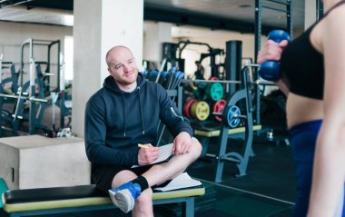 Beden eğitimi hocası not defterine genç atletik sarışının spor salonunda elinde dambıllarla egzersiz yapmasının sonuçlarını yazar.