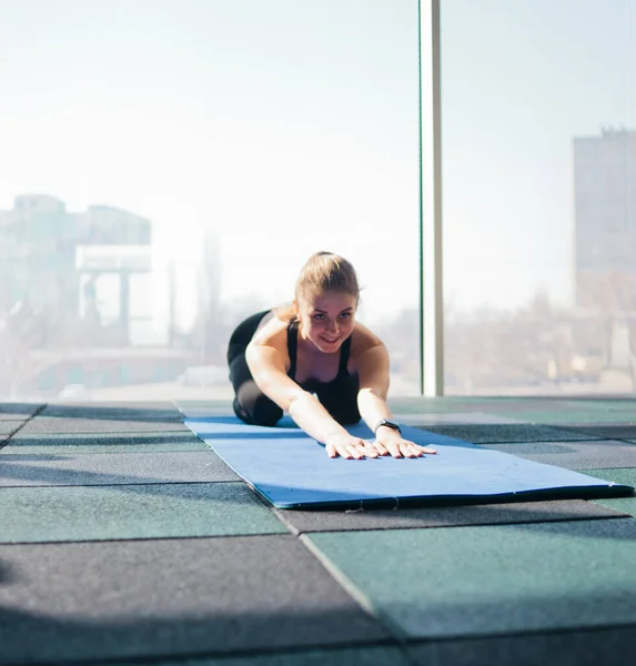 Young woman doing stretching for the whole body on yoga mat against the background of large panoramic window in the gym. Yoga training