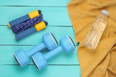 Düz yatış tarzı spor konsepti. Dambıllar, ip atlama, havluyla su şişesi. Mavi ahşap arka planda spor aletleri. Üst görünüm