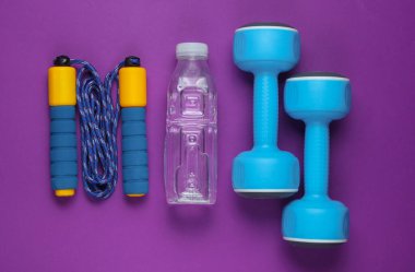 Flat lay style sport concept. Dumbbells, jump rope, bottle of water. Sports equipment on purple background. Top view clipart
