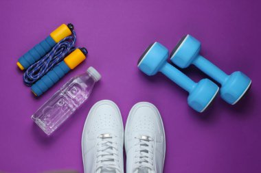 Flat lay style sport concept. Dumbbells, sneakers, jump rope, bottle of water. Sports equipment on purple background. Top view clipart