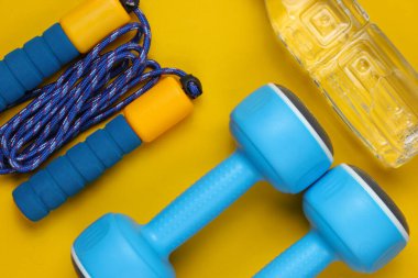 Düz yatış tarzı spor konsepti. Dambıllar, ip atlama, bir şişe su. Sarı arka planda spor malzemeleri. Üst görünüm