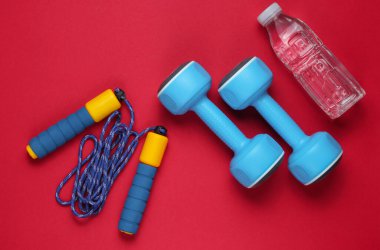 Düz yatış tarzı spor konsepti. Dambıllar, ip atlama, bir şişe su. Kırmızı arka planda spor malzemeleri. Üst görünüm