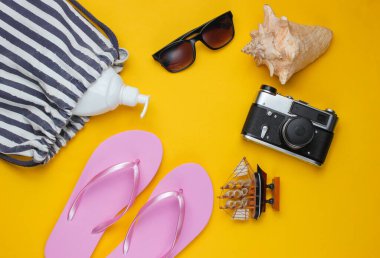 Yaz hala hayattır. Plaj aksesuarları. Pembe terlikler, çanta, retro kamera, güneş kremi şişesi, güneş gözlüğü, sarı kağıtta deniz kabuğu. Düz yatıyordu. Uzayı kopyala Üst görünüm