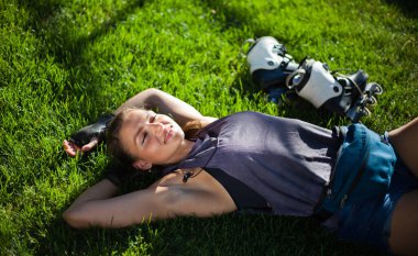 Genç sporcu kadın patenci parkta çimlerin üzerinde dinleniyor ve kulaklıkla müzik dinliyor.